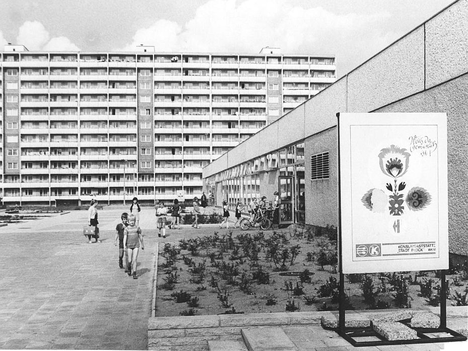 Schulkinder gehen in ein flaches Gebäude. Im Hintergrund steht ein elfgeschossiger Plattenbau.