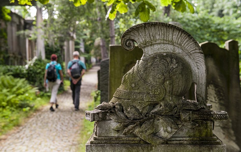 An einer Steinplastik auf einem Friedhof, die wie ein Helm aussieht, gehen ein Mann und eine Frau vorbei.