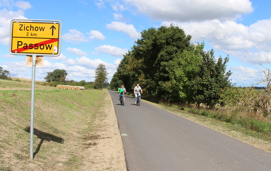 Foto: asphaltierter Weg mit 2 Radfahrern und dem Ortseingangsschild von Passow