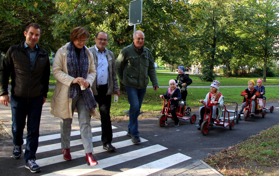 Foto: 4 Erwachsene überqueren einen Zebrastreifen, 5 Kinder warten mit ihren Fahrzeugen