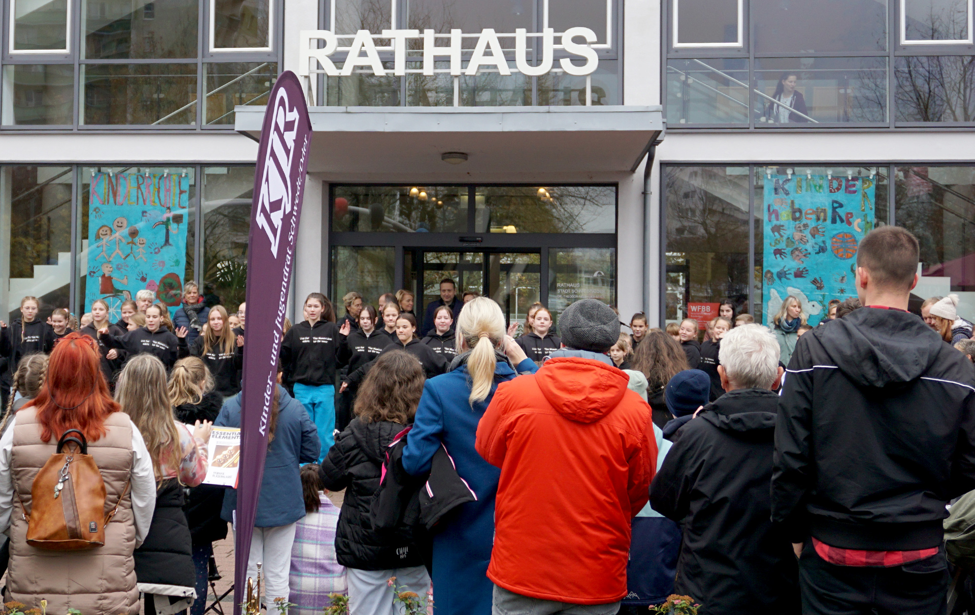 Foto: Fahnen in der Rathausfensterfront, davor Publikum der Aktion