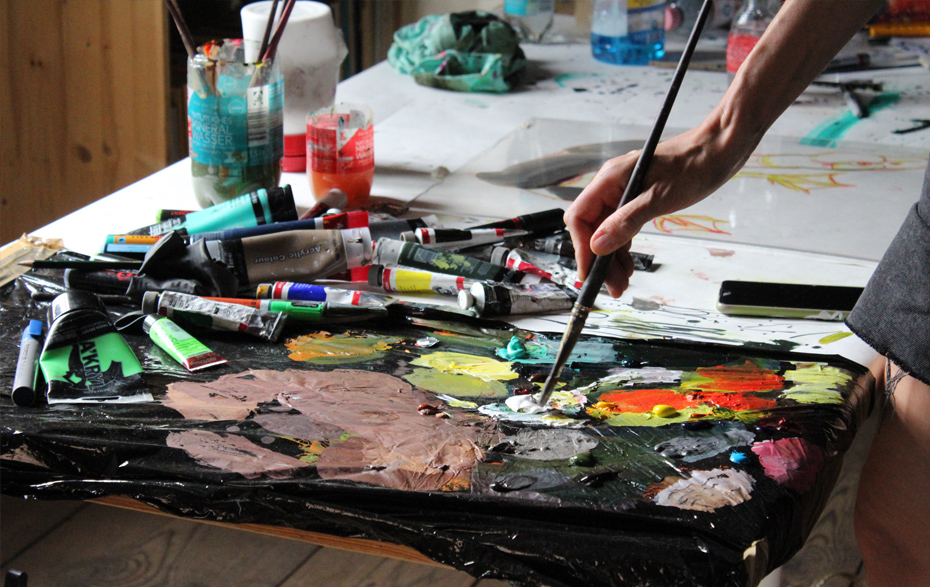 Foto: Hand mit Pinsel taucht in die Palette mit Farben, daneben Farbtuben und Wasserbehälter 