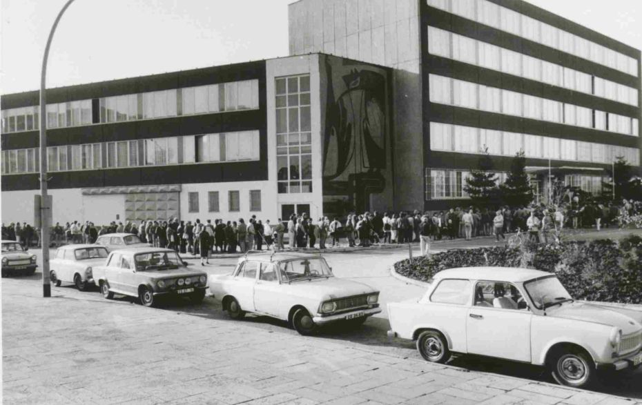 Blick auf eine Straße mit Autos. Im Hintergrund ein Gebäude mit einer langen Menschenschlange.