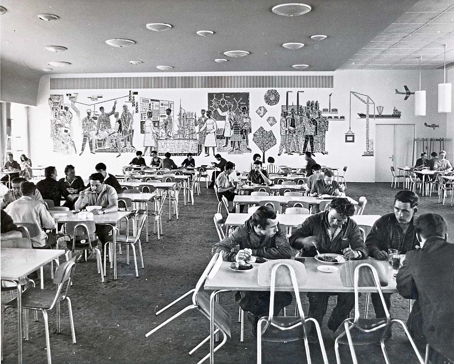 Sitzgruppen mit Menschen beim essen. im Hintergrund ein Wandbild, was diie Geschichte des Erdölverarbeitswerkes Schwedt zeigt.