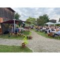 Unter einem Dach stehen Frau Felsberg und Frau Stockfisch im Garten des Museums und sprechen an Mikrofon. Vor der Überdachung sitzen Menschen auf Stühlen. Daneben stehen Blumensträuße bereit. 