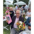 Wolfgang Lichtenberg sitzt und hält in einer Hand ein rosa Buch und in der zweiten einen Blumenstrauß. Hinter ihm sitzen Menschen im Garten. 