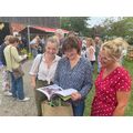 Frauen lachen mit einem Buch in der Hand. Hinten stehen viele Menschen bei einem Fest im Garten.