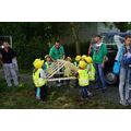 Foto: Kinder mit Bauhelmen und gelben Westen schauen zwei Männern tragen einen Holzdachstuhl.