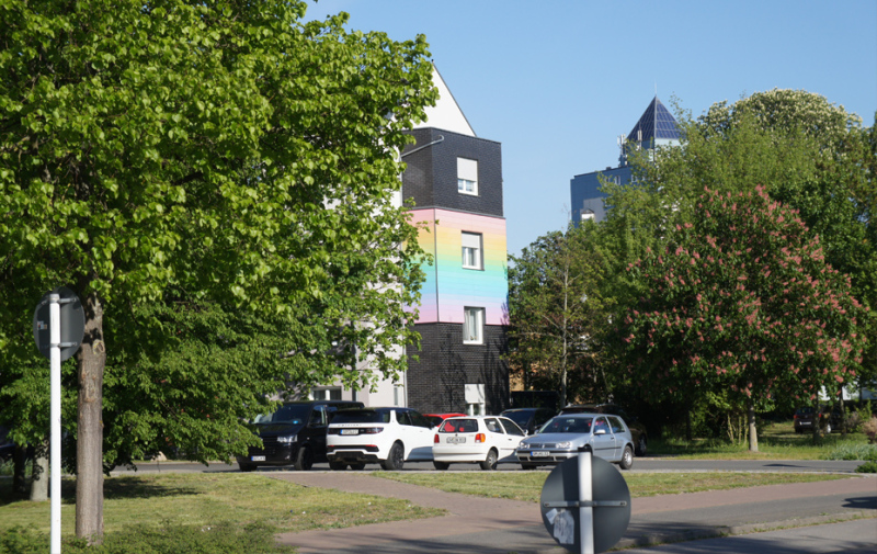 Foto: parkende Autos neben dem Wohnhaus mit den Regenbogenfarben und viele grüne Bäume