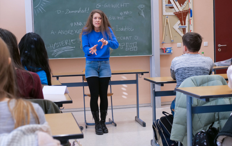 Foto: Schauspielerin steht gestikulierend vor einer Schultafel