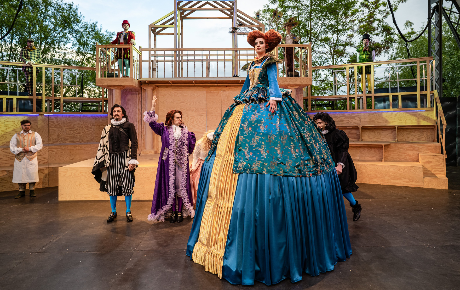 Foto: Schauspielerin in einem üppigen riesigen Kostüm umgeben von weiteren Schauspielen in Kostüm auf der Open-Air-Bühne